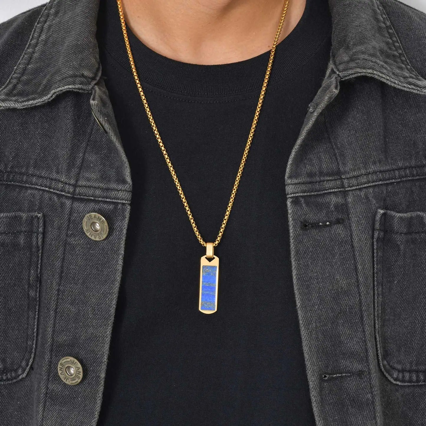 Person wearing a gold necklace with a blue pendant against a black shirt and denim jacket.