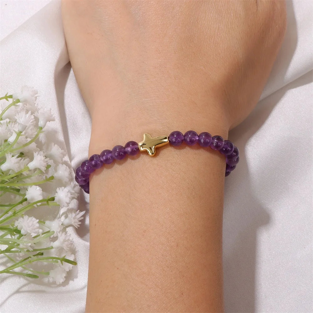 Purple beaded bracelet with a gold cross charm on a wrist, with white flowers in the background.