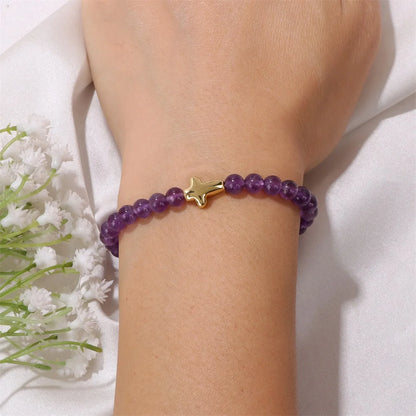 Purple beaded bracelet with a gold cross charm on a wrist, with white flowers in the background.