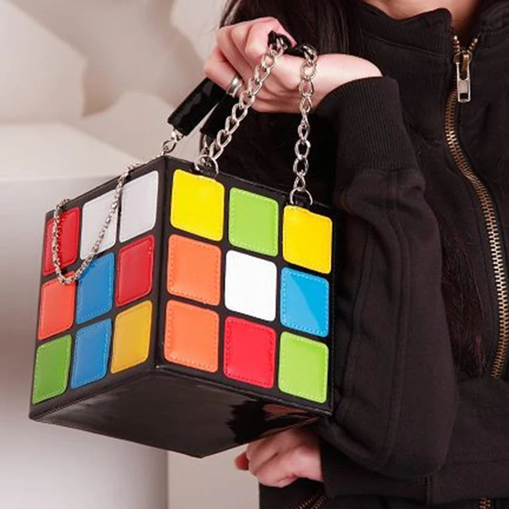 Colourful cube-shaped handbag with chain handle held by a person wearing a black jacket.