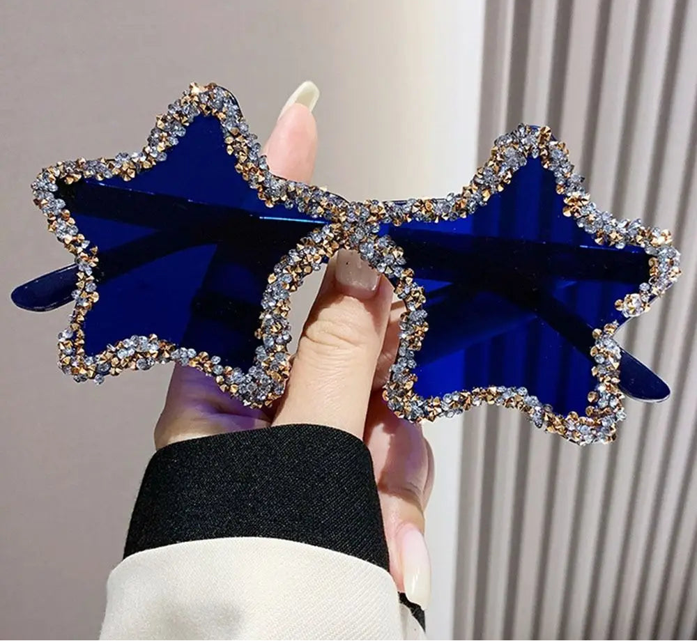 Blue star-shaped sunglasses with rhinestone details held by a hand against a neutral background.