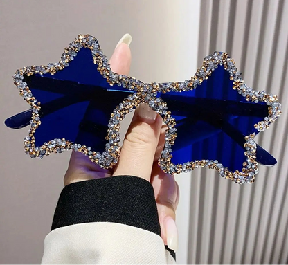 Blue star-shaped sunglasses with rhinestone details held by a hand against a neutral background.