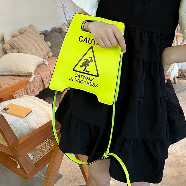 Person holding a bright yellow sign with 'CAUTION CATWALK IN PROGRESS' text, wearing a black dress.