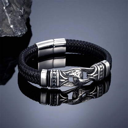 A black braided leather bracelet with a polished silver-toned metal clasp featuring an engraved fish design, displayed on a reflective dark surface.