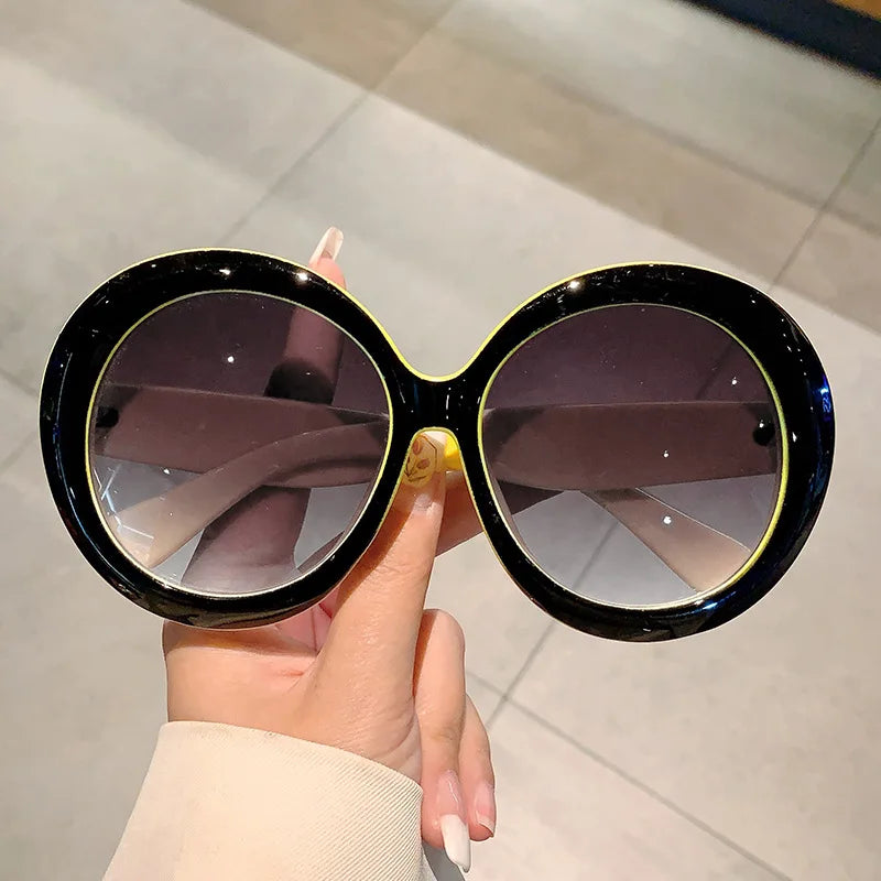 Round sunglasses with black frame and yellow accents held by a hand on a light background