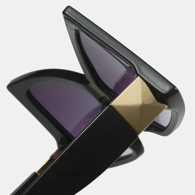 Close-up detail shot of black sunglasses with dark purple-tinted lenses and a metallic gold geometric frame accent, shown on a light grey background.