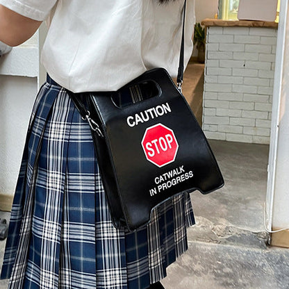 Person wearing a white shirt and plaid skirt holding a black bag with 'Caution: Catwalk in Progress' text and symbol sign in an indoor setting.