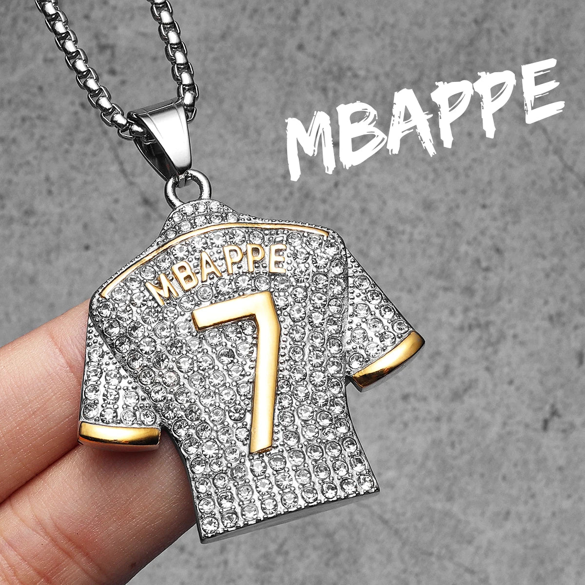 Pendant shaped like a diamond-encrusted sports jersey with 'MBAPPE' and number '7', held by a hand against a grey background.