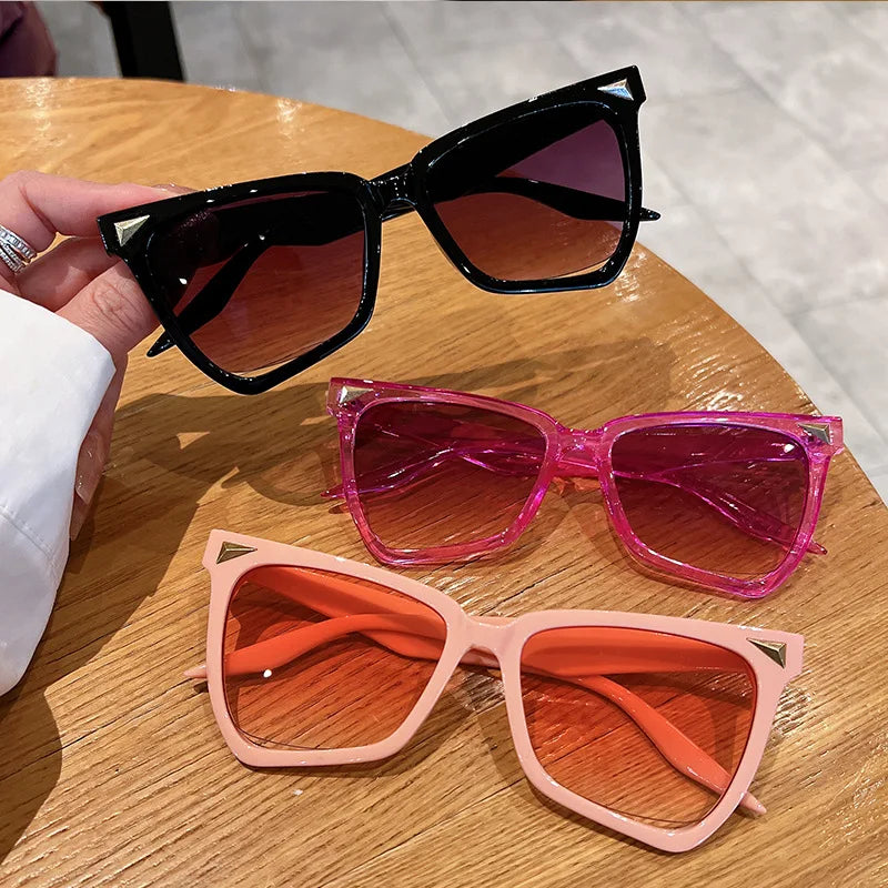 Three pairs of cat-eye sunglasses in black, pink, and orange on a wooden surface.