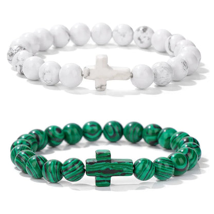 Two beaded bracelets, one white with a cross charm and the other green with a cross charm, on a white background.