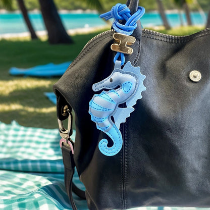 A black leather shoulder bag with a blue seahorse-shaped keychain charm, photographed outdoors on a beach blanket with a blurred tropical beach background.