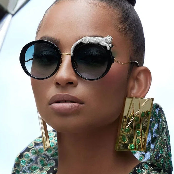 Woman wearing large round sunglasses and gold earrings with green gemstones.