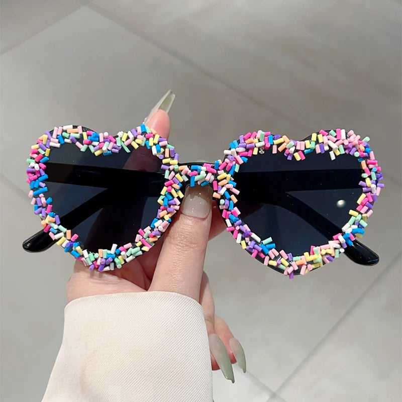 Heart-shaped sunglasses with colourful sprinkled frame held by a hand against a neutral background