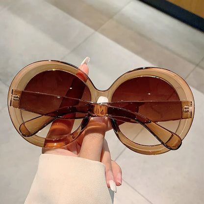 Large round sunglasses with brown lenses held by a hand against a neutral background