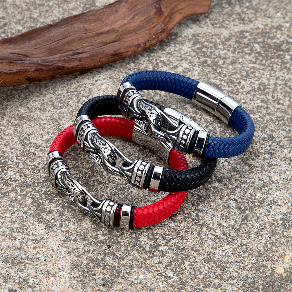 Three braided leather bracelets with ornate silver-toned metal links are arranged on a textured stone surface. One bracelet is blue, one is red, and one is black, each featuring a large decorative silver clasp.