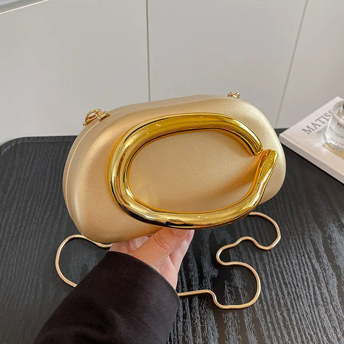 Gold clutch bag held by a hand on a dark surface with a light grey wall in the background.