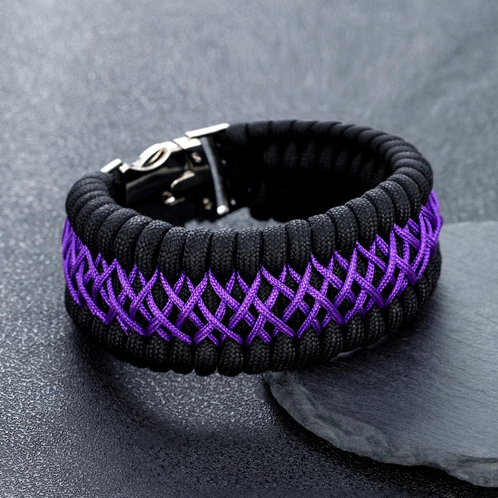 A black paracord bracelet with a purple woven pattern and a silver metal clasp, resting on a dark grey textured surface.