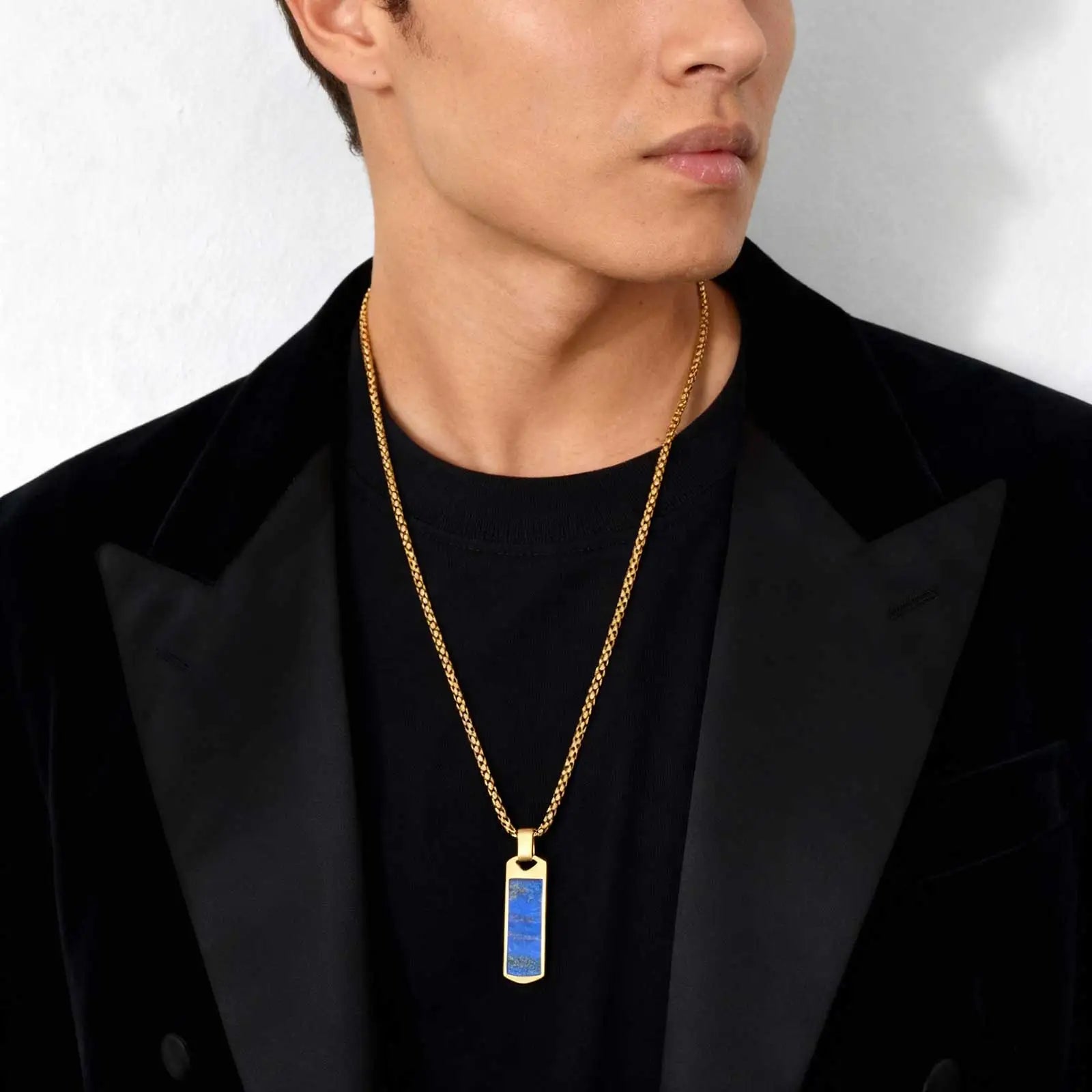 Man wearing a black jacket with a gold necklace featuring a blue pendant.