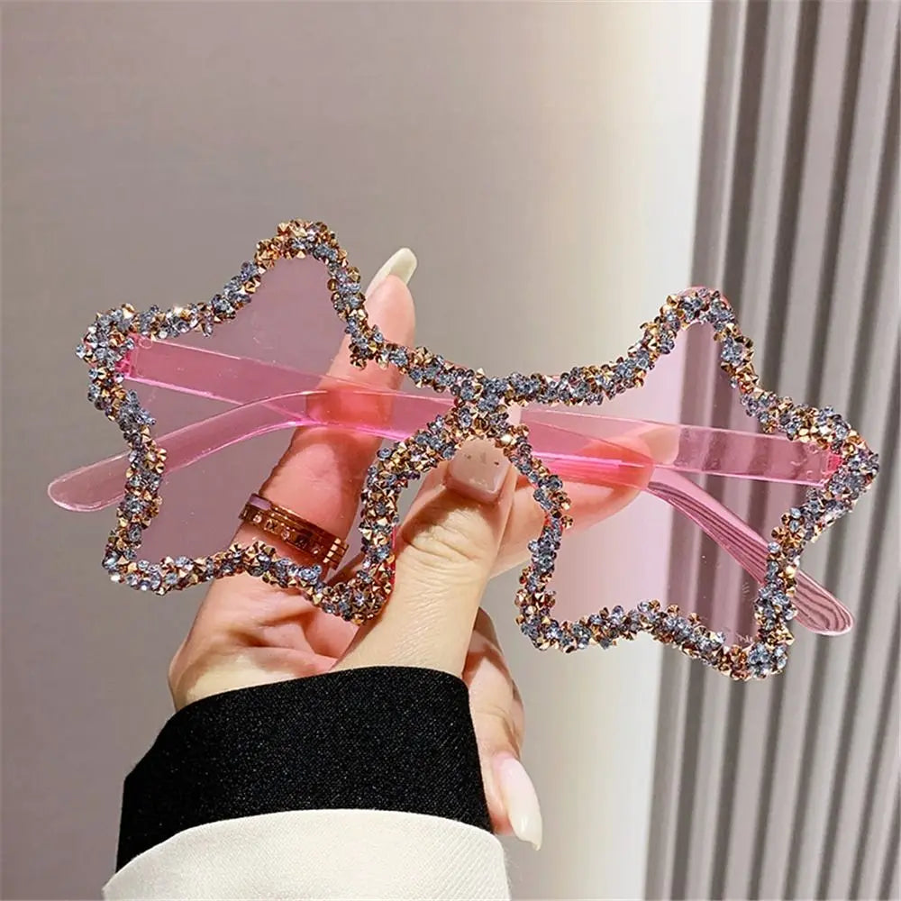 Pink Star-shaped sunglasses with rhinestone details held by a hand against a neutral background