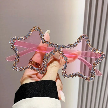 Pink Star-shaped sunglasses with rhinestone details held by a hand against a neutral background