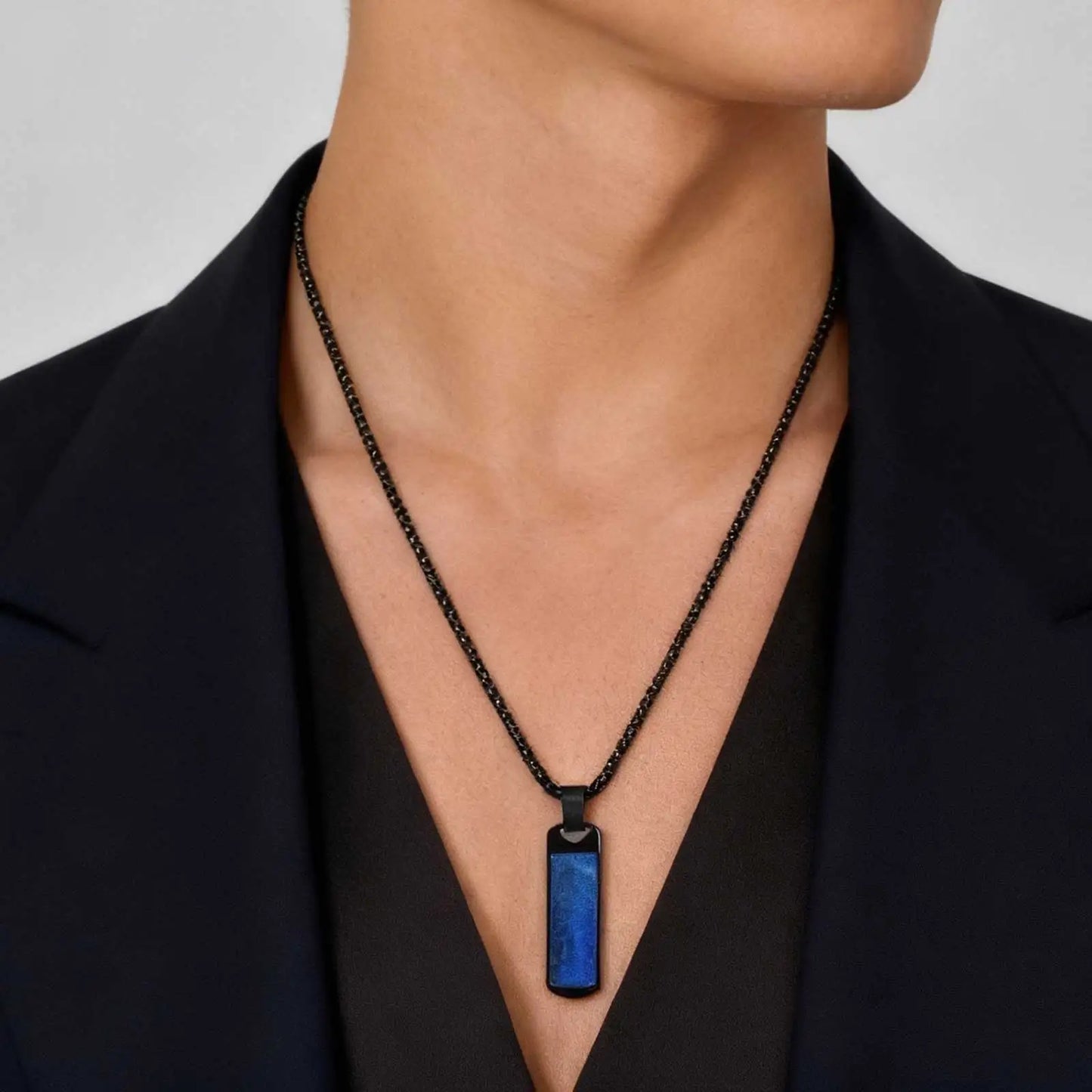Person wearing a black chain necklace with a blue pendant against a neutral background