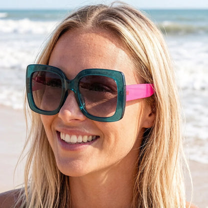 A smiling blonde woman on a beach wearing oversized square sunglasses with translucent teal-blue frames and bright pink temple arms.