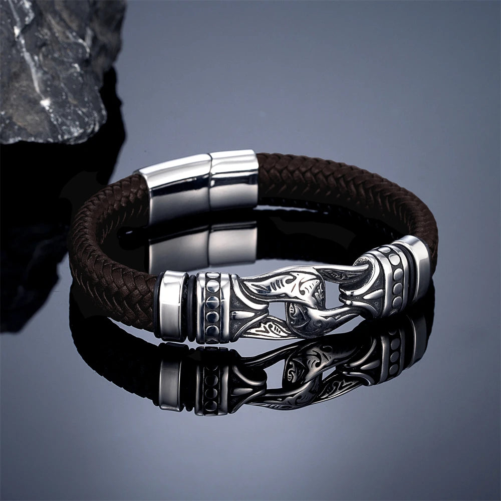A black braided leather bracelet with a polished silver-toned metal clasp featuring an engraved fish design, displayed on a reflective dark surface.