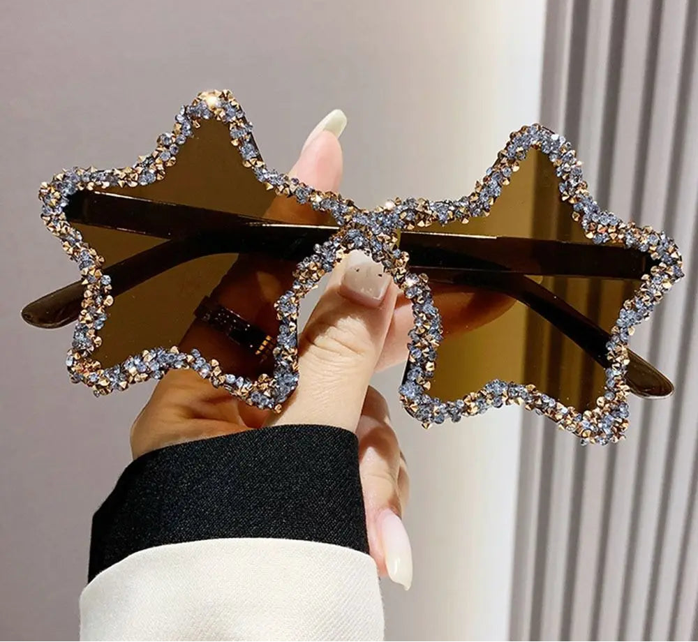 Star-shaped sunglasses with decorative frame held by a hand against a neutral background