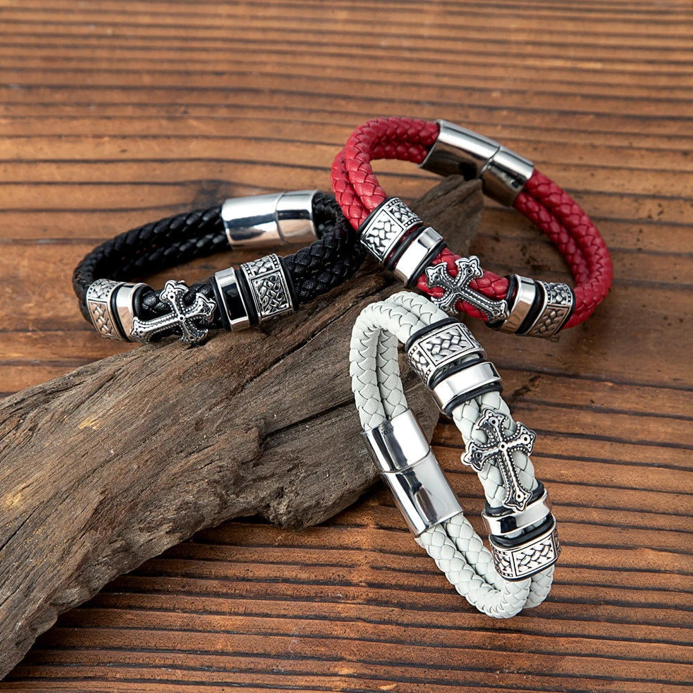 Three braided leather bracelets with silver-toned cross and decorative bead details, arranged on a rustic wood surface. One bracelet is black, one is red, and one is light grey.