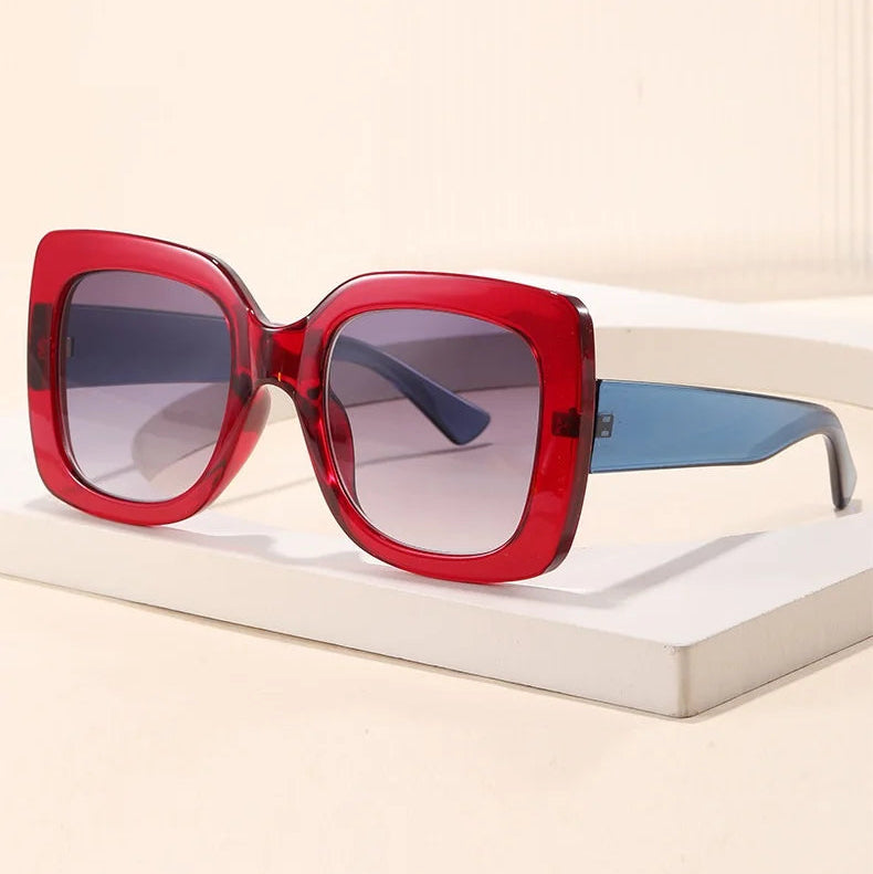 Oversized square sunglasses with a translucent red frame, gradient grey lenses, and light blue temple arms, displayed on a white book against a beige background.