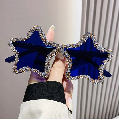 Blue star-shaped sunglasses with rhinestone details held by a hand against a neutral background
