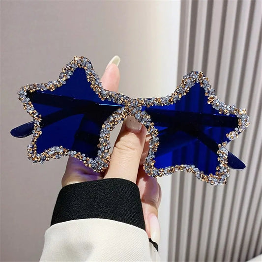 Blue star-shaped sunglasses with rhinestone details held by a hand against a neutral background