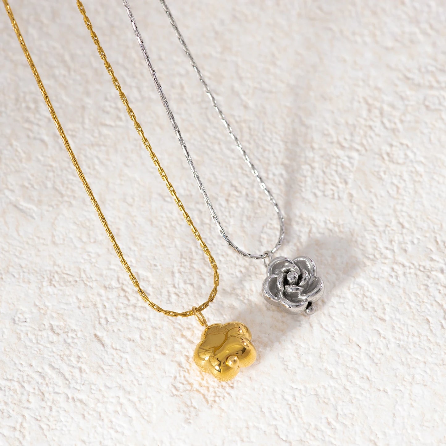 Two necklaces with flower pendants, one gold and one silver, on a textured white background.