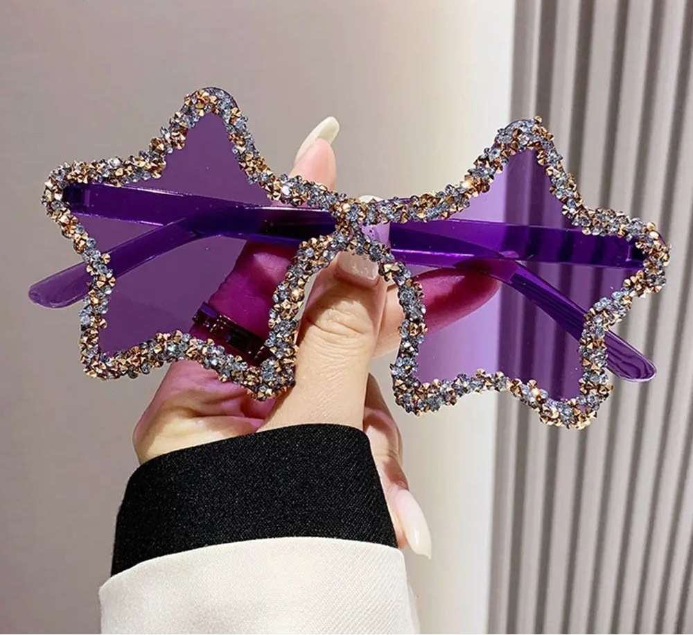 Purple star-shaped sunglasses with rhinestone details held by a hand against a neutral background.