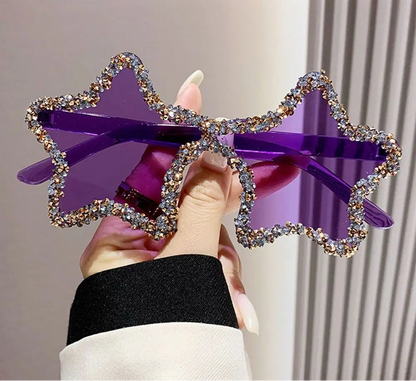 Purple star-shaped sunglasses with rhinestone details held by a hand against a neutral background.