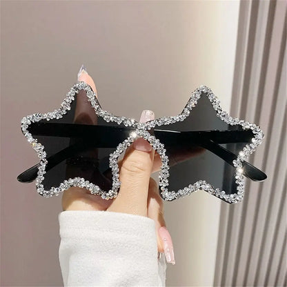 Star-shaped sunglasses with rhinestone frame held by a hand against a neutral background