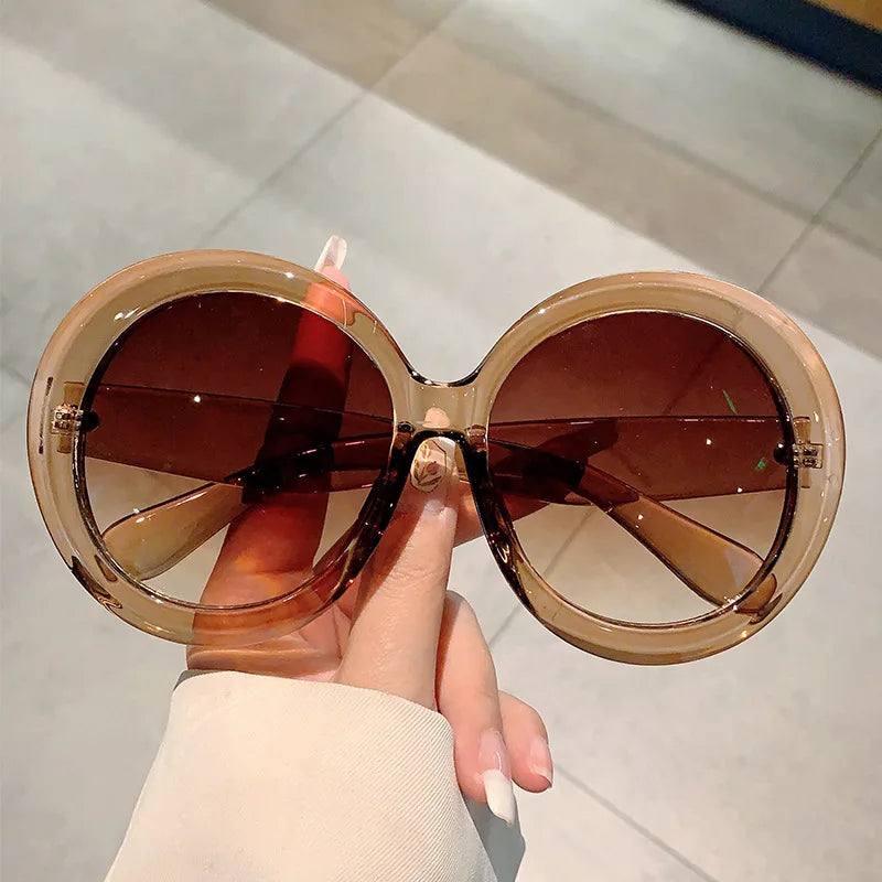 Large round sunglasses with brown lenses held by a hand against a light background