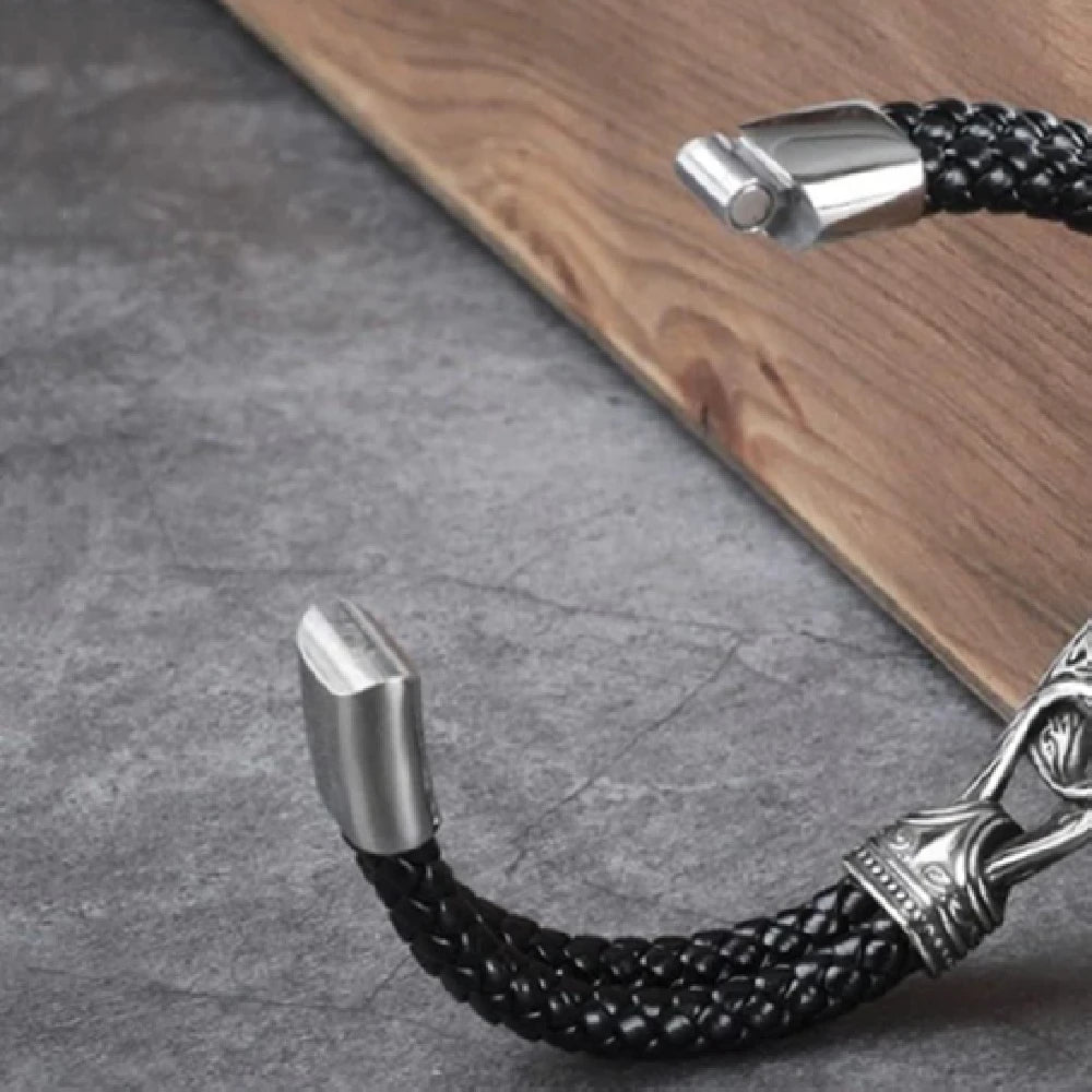 A close-up of a black braided leather bracelet with polished silver-tone metal ends, shown resting on a grey stone-like surface next to a wooden board.