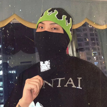 Person wearing a black balaclava with a green and black beanie, standing in front of a cityscape at night.