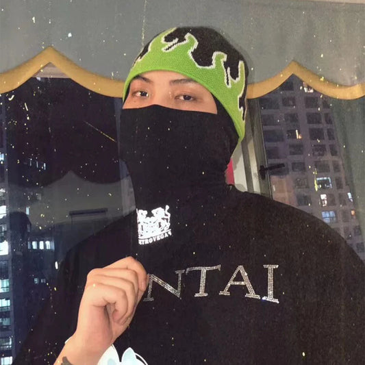 Person wearing a black balaclava with a green and black beanie, standing in front of a cityscape at night.