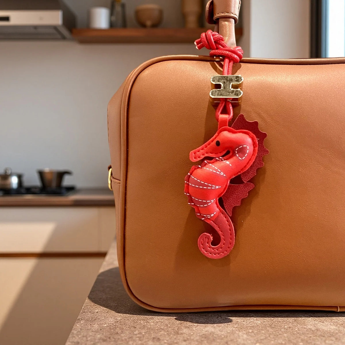 A tan leather bag with a red seahorse-shaped hanging charm and a red knotted strap detail, photographed on a countertop in a kitchen setting.