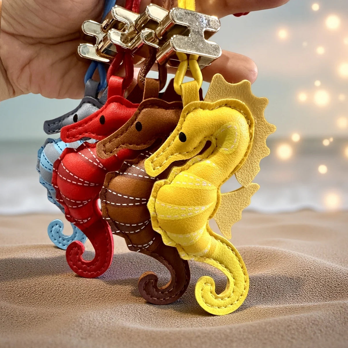 A hand holds a set of four small seahorse-shaped keychains or bag charms in different colours: yellow, red, brown, and blue. The keychains have a stitched leather or faux-leather finish with white contrast stitching and gold-tone metal key rings, shown against a soft beach background with warm bokeh lights.