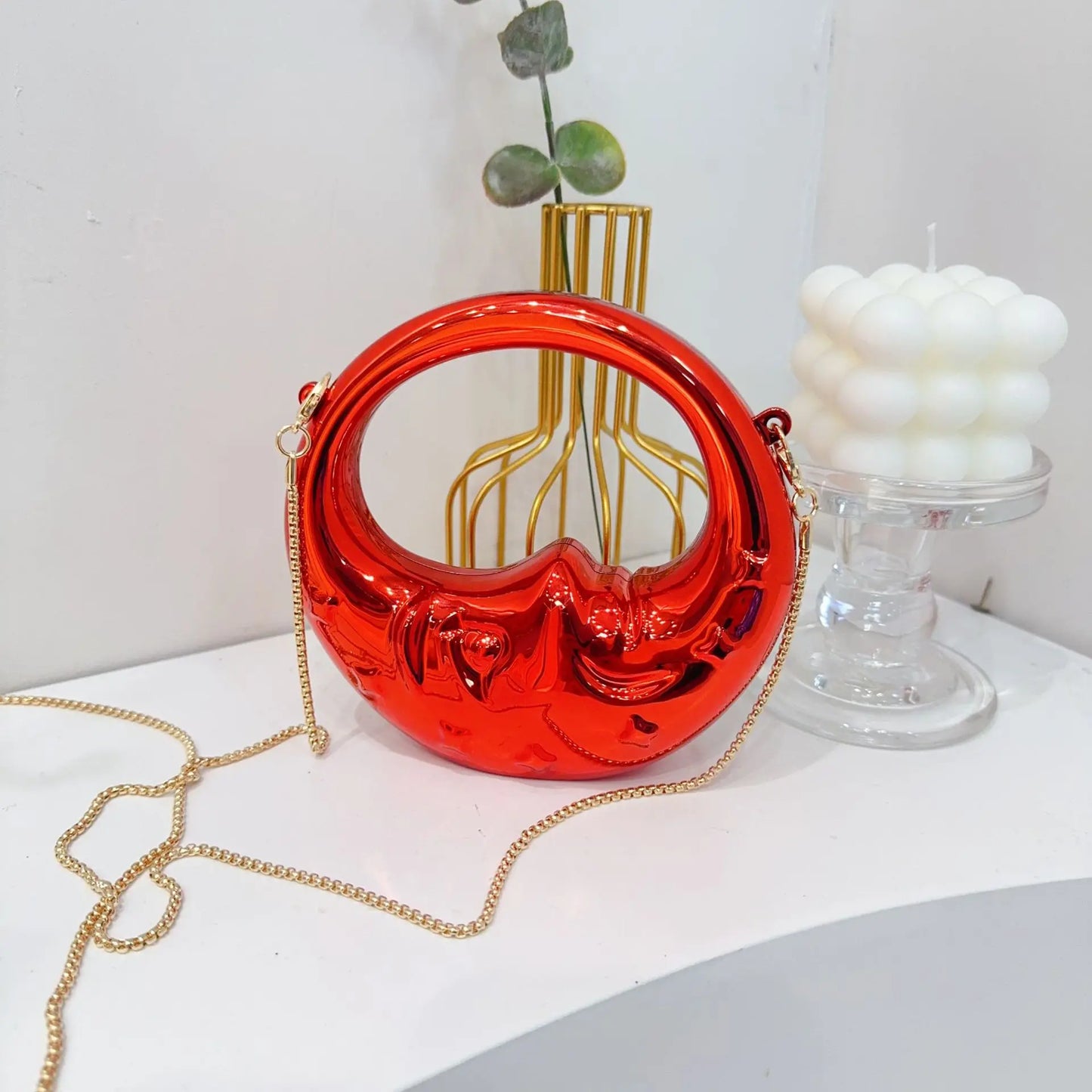 Red crescent handbag with gold chain on a white surface with decorative items.