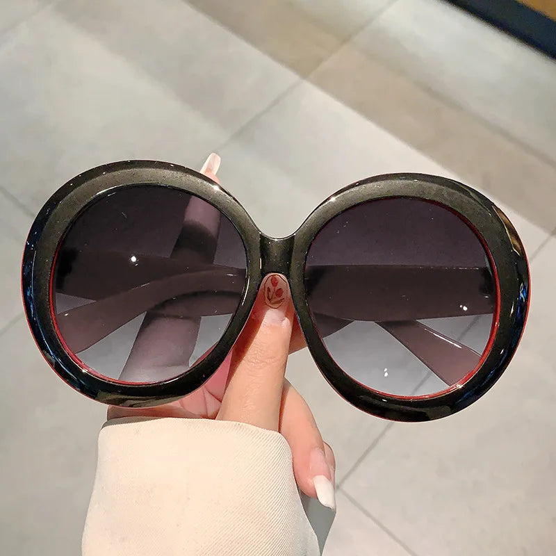 Round sunglasses with black and red accents frame and dark lenses held by a hand against a light background