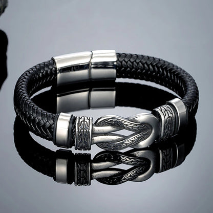 A black braided leather bracelet with a polished silver-toned metal clasp featuring a stylized knot design, displayed on a reflective grey surface.
