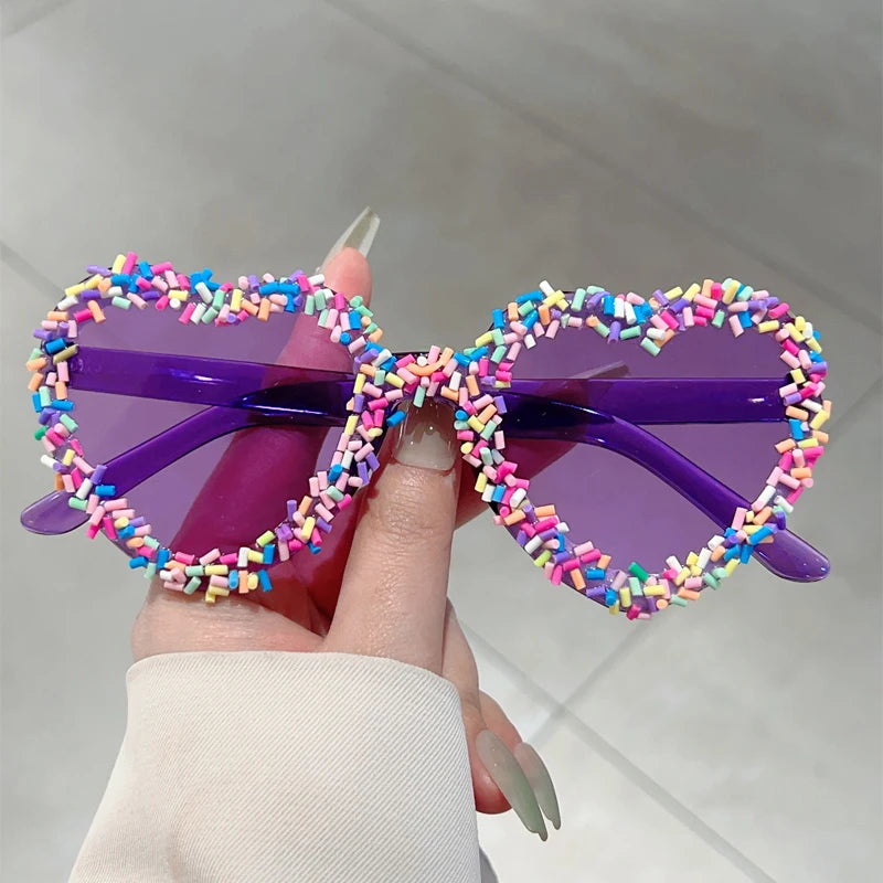Colourful heart-shaped sunglasses with sprinkles held by a hand against a neutral background