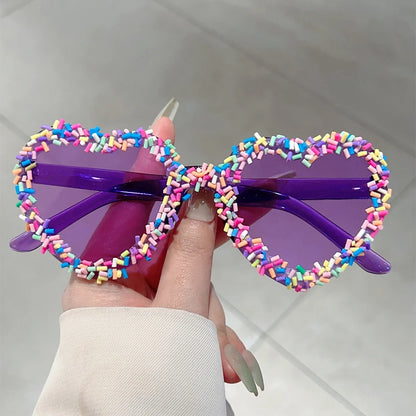 Colourful heart-shaped sunglasses with sprinkles held by a hand against a neutral background