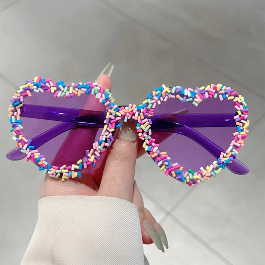 Colourful heart-shaped sunglasses with sprinkles held by a hand against a neutral background