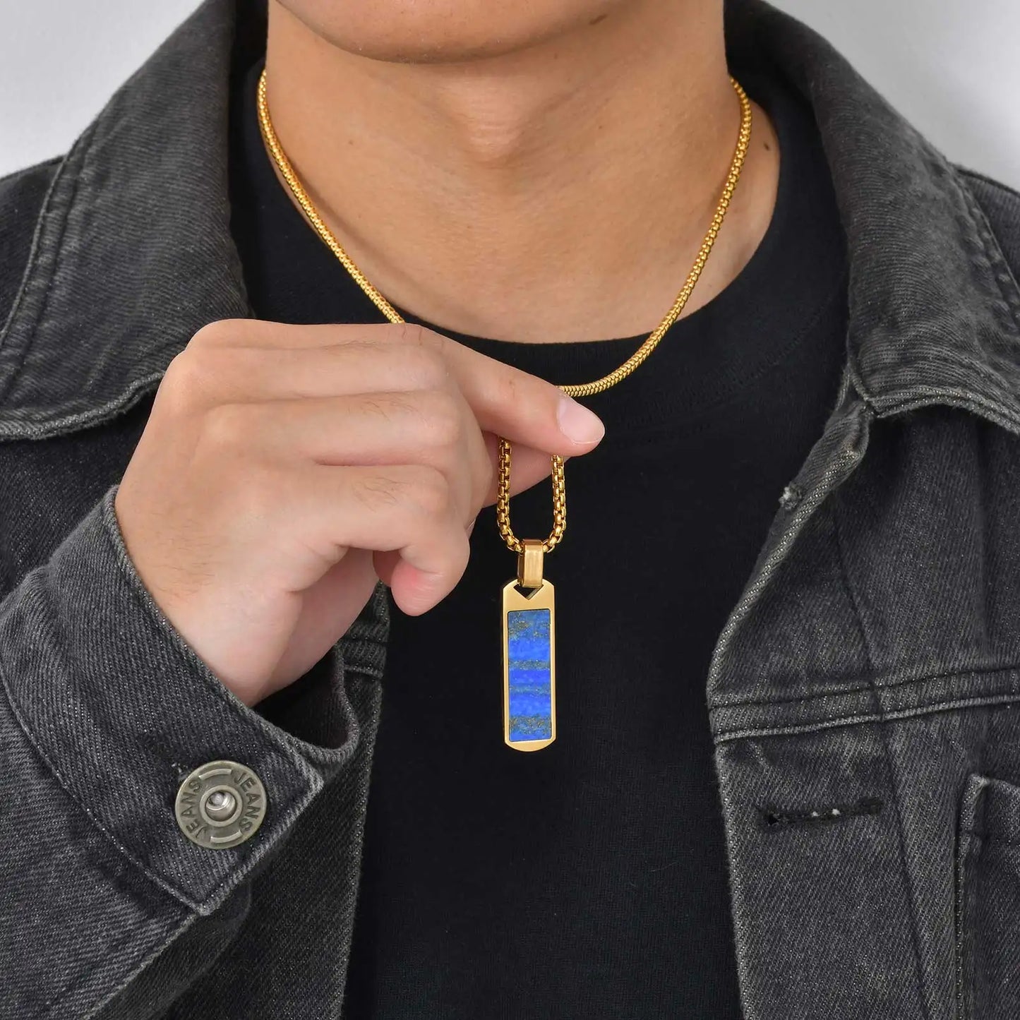 Person wearing a gold necklace with a blue lapis lazuli pendant, pointing to it.