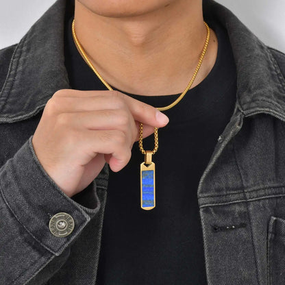 Person wearing a gold necklace with a blue lapis lazuli pendant, pointing to it.